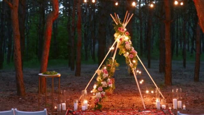 波西米亚风格的tipi木制拱门装饰着燃烧的蜡烛，玫瑰和pampass草，晚上在松树林的户外婚礼场地上