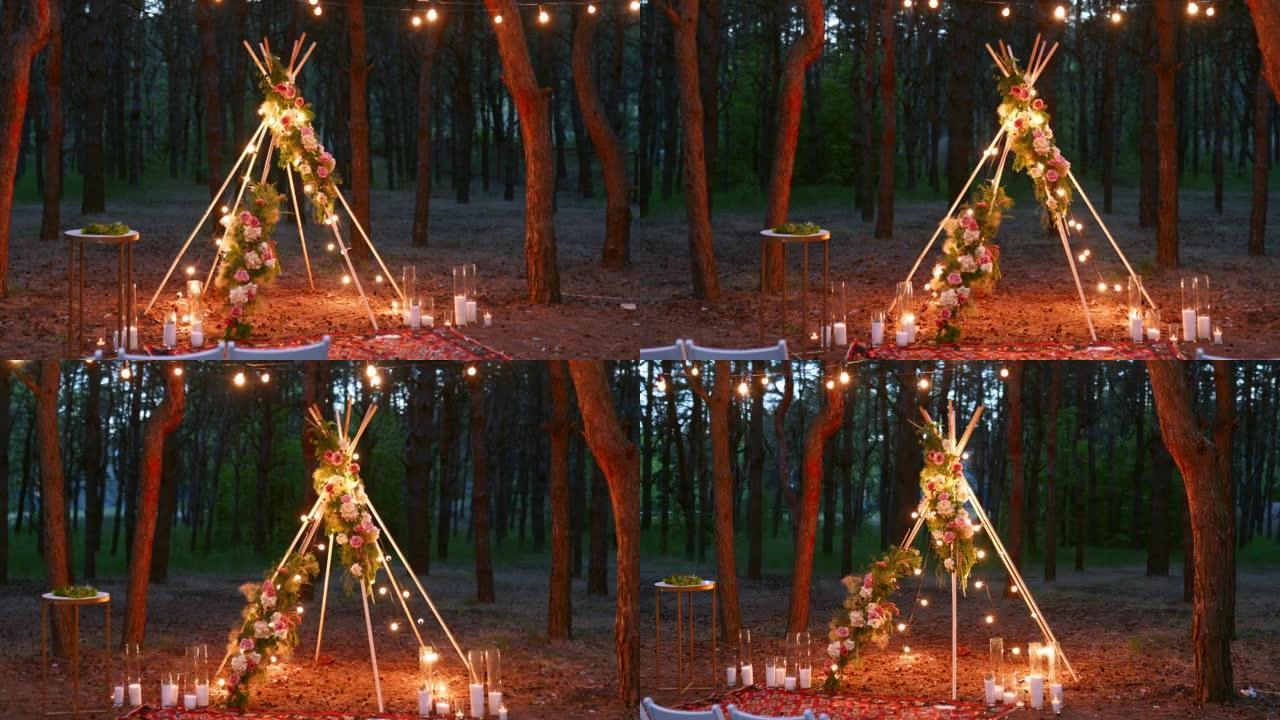 波西米亚风格的tipi木制拱门装饰着燃烧的蜡烛，玫瑰和pampass草，晚上在松树林的户外婚礼场地上