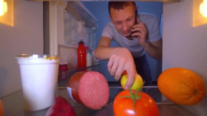 男人正在打电话，打开冰箱，拿出一个苹果和西红柿。