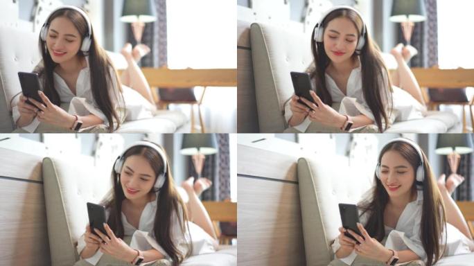 美丽的亚洲女人穿着性感的连衣裙享受在客厅沙发上戴着耳机在智能手机技术上听音乐