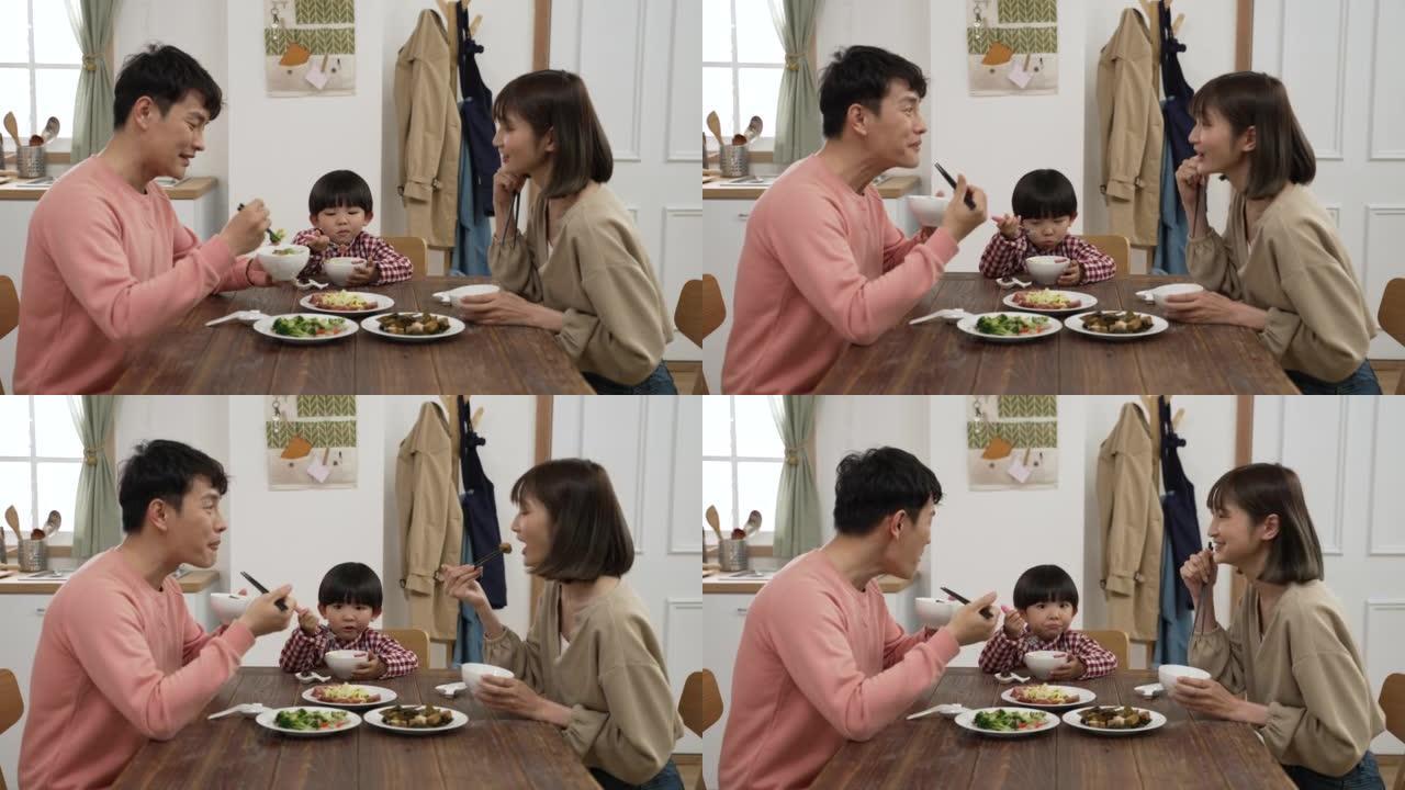 快乐的亚洲三口之家在餐厅共进午餐。微笑的母亲请父亲用手势尝试蔬菜菜