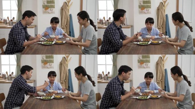 快乐的亚洲父亲、母亲和学龄前男孩在家一起吃午饭。这对恩爱的夫妇互相给食物和儿子的碗