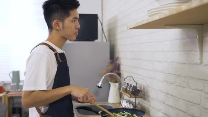 侧面肖像亚洲年轻丈夫做饭午餐正在点头，同时在公寓的厨房炉灶上尝试他的just passage蔬菜。