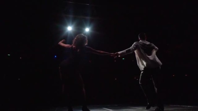 在泛光灯光的舞台上，一群交谊舞舞者在黑暗中表演着拉丁美洲的古典舞步，摆动着臀部和手臂，镜头从下面掠过