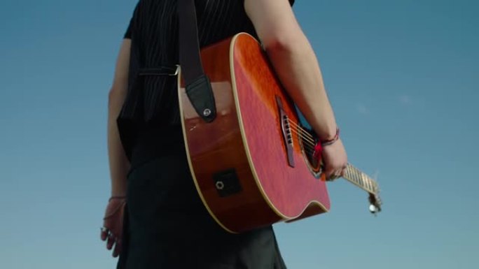 年轻音乐家摇滚男孩拿着木制原声吉他走路的仰视图。时髦的男流浪歌手打扮得像嬉皮士，带着乐器散步。特写蓝