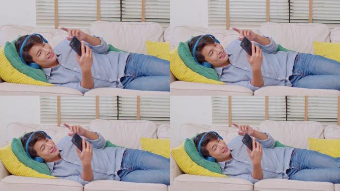 亚洲帅哥躺在沙发上戴着耳机听音乐，用手机或智能手机在家在线选择音乐应用或视频媒体。