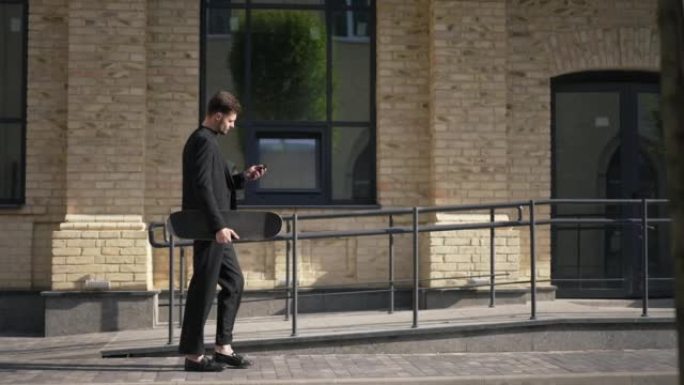 侧视图严肃成功的年轻商人带着滑板和智能手机在城市街道上的阳光下行走。现场摄像机跟随白人优雅的男人在阳