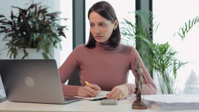 她办公室的一位女律师正在笔记本电脑上工作，并在笔记本上做笔记。一名欧洲公证妇女在笔记本电脑上打字