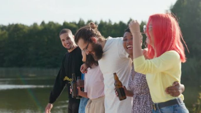 朋友在湖边相聚一起度过时光，他们拥抱着彼此向前走，笑着，胡闹着手里拿着啤酒瓶，头发被风吹起