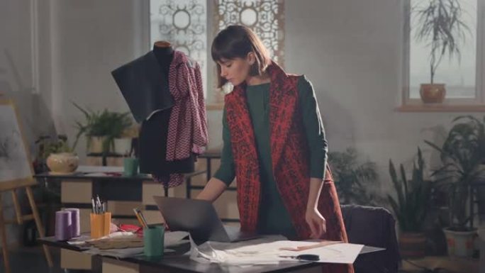在工作室工作的年轻女孩时装设计师。她正站在桌子旁，手提电脑和素描。