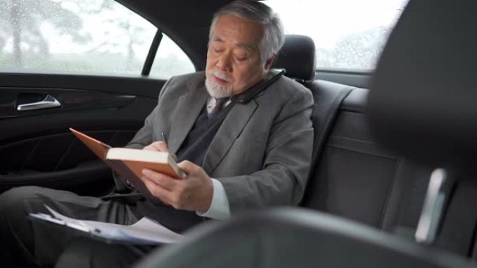 4k亚洲高级商人坐在汽车后座上，在电话交谈时在书中写商业计划书
