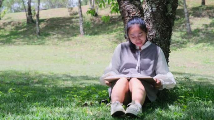 亚洲妇女坐在树下在公园看书。