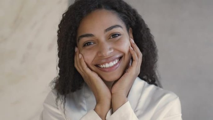自然美概念。年轻的积极的非洲裔美国女士对着镜头微笑，抚摸着她光滑的脸颊的特写镜头