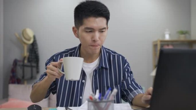 在计算机上在家工作的有吸引力的亚洲商人正在从杯子里吞茶，同时在后台的客厅检查财务文件