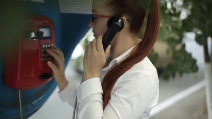 一位年轻的女商人穿着白色上衣，戴着红头发，戴着眼镜，在电话亭上打电话到另一个国家。