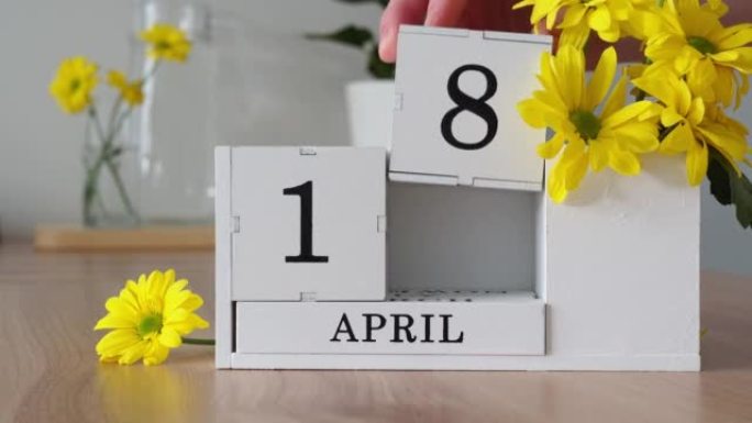 春季月份4月18日。女人的手翻过一个立方历法。黄色花朵旁边的桌子上的白色万年历。在一个月内更改日期。
