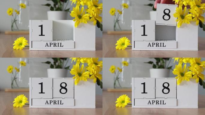 春季月份4月18日。女人的手翻过一个立方历法。黄色花朵旁边的桌子上的白色万年历。在一个月内更改日期。