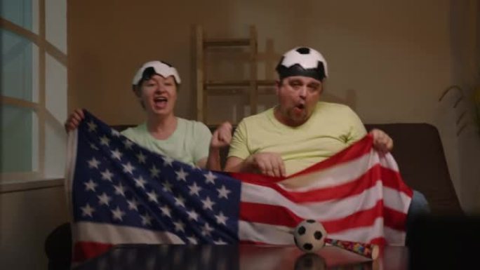 坐在沙发上的足球迷在家里的电视上欢呼挥舞着美国国旗观看比赛