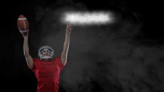 男性橄榄球运动员拿着球指着体育场灯光，黑色背景上的烟雾
