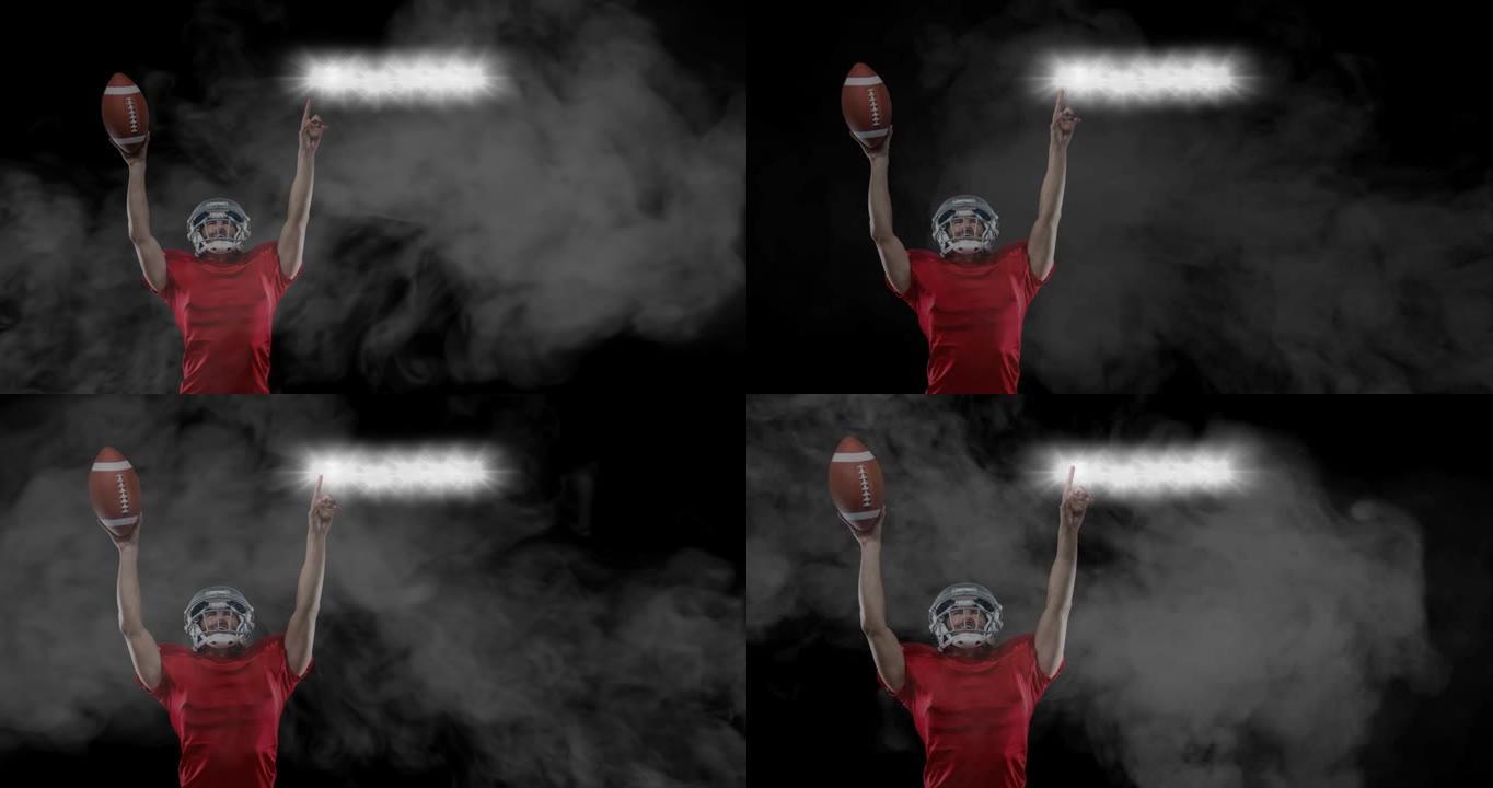男性橄榄球运动员拿着球指着体育场灯光，黑色背景上的烟雾