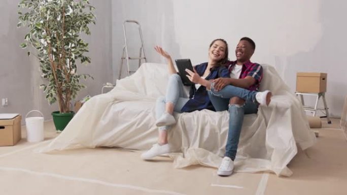 不同国籍的夫妇坐在客厅的沙发上，在装修期间新买的公寓里，情侣在平板电脑上讨论装饰家具墙壁颜色观看项目
