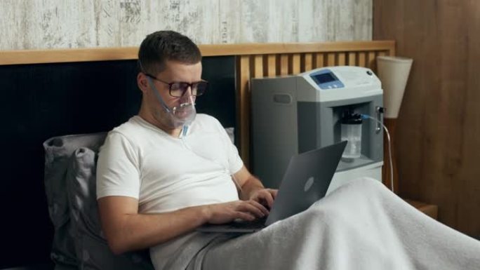 一个生病的年轻人的肖像使用氧气面罩和氧气浓缩器在家中治疗并在笔记本电脑上工作。