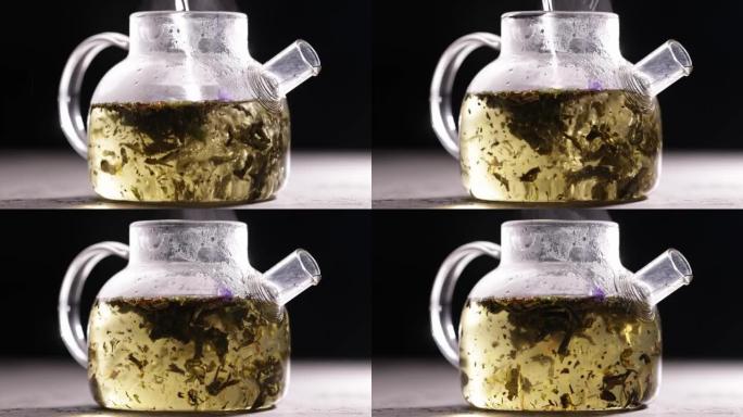 在深色背景下的玻璃透明茶壶中酿造绿茶的过程。将热水倒入带茶叶的水壶中。