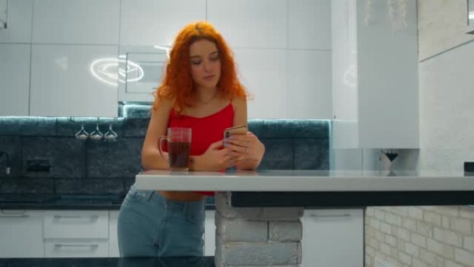 红头发的年轻女子在厨房里喝茶，透过手机看