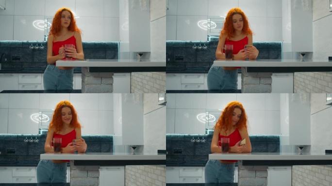 红头发的年轻女子在厨房里喝茶，透过手机看
