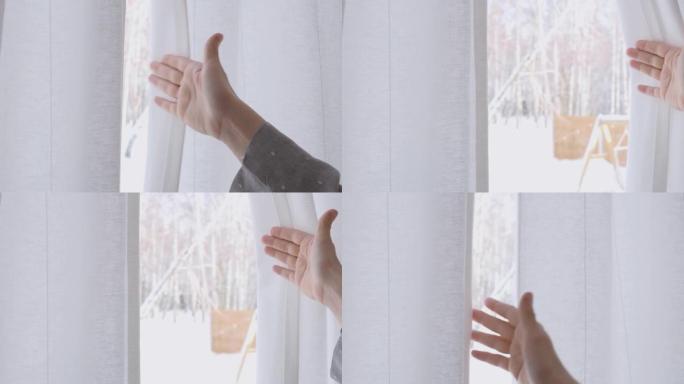 女人的手伸手去拿窗帘，打开窗帘，看着窗外，窗外阳光明媚。冬季景观和被雪覆盖的森林。在窗前等待的女人