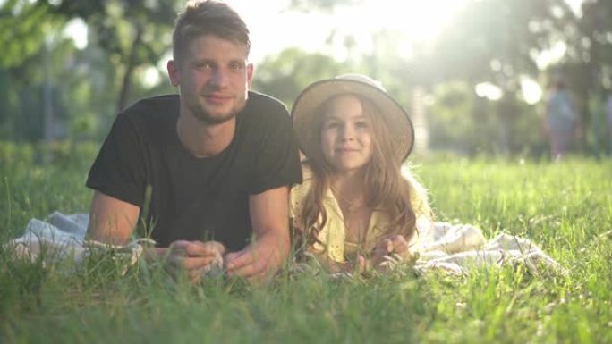 正面看积极的年轻父亲和小女儿躺在阳光下的公园草地上微笑着说话。轻松积极的高加索女孩和男人在阳光中享受