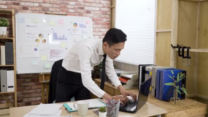 一名亚洲商人站在办公桌前，在一间现代化的阁楼办公室里，在日光下阅读完纸质文件上的数据后，正俯身在笔记