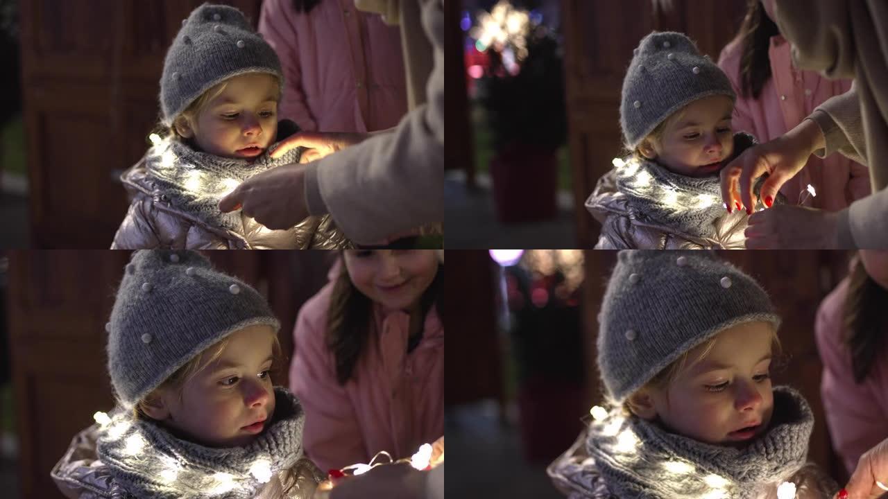 好奇的蹒跚学步的女孩观察她的母亲是如何把圣诞灯放在围巾上的