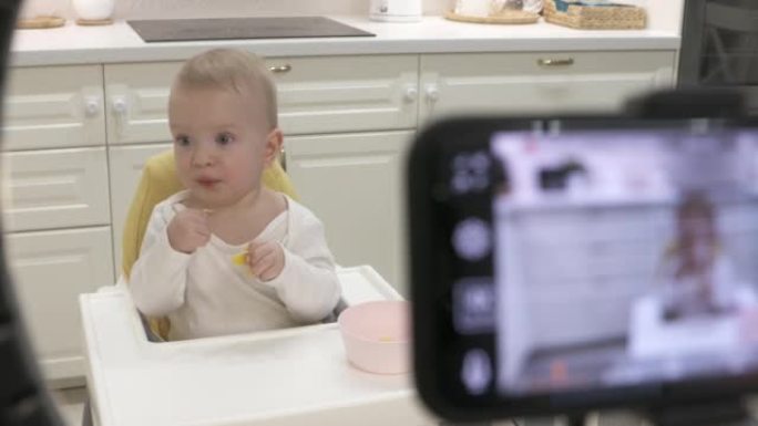 父母在家里高脚椅上的孩子的智能手机上录制视频，用手机和铃声录制一岁大的男婴水果的照片视频。