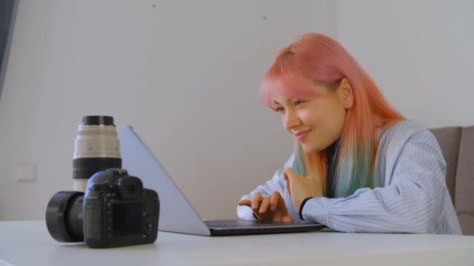 摄影师女人与彩色彩虹头发在笔记本电脑上打字文本，带有有趣的微笑表情。年轻的乌克兰女性与客户在线交流