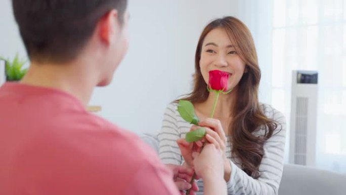 亚洲年轻人惊喜并将玫瑰花送给美丽的女友。迷人的浪漫新婚夫妇男女花时间在家里一起庆祝周年纪念日和情人节