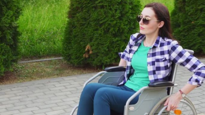 迷人的残疾女孩穿着衬衫和眼镜坐在轮椅上，在公园里慢悠悠地走着