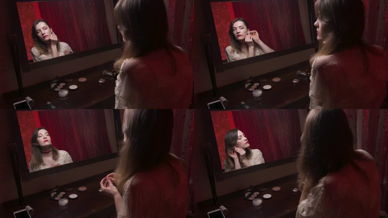 漂亮的白人年轻女子在化妆镜前的卧室里穿石头耳环。昂贵的红光下的闺房摄影。迷人的女孩去舞会