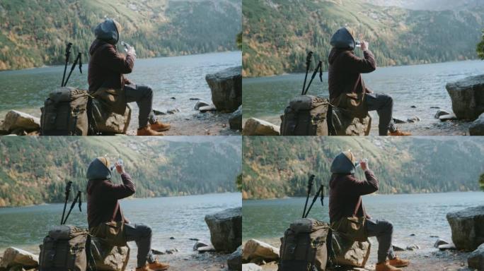 男性徒步旅行者背包客在高地艰苦跋涉后，坐在美丽的山湖岸上的石头上，从旅游酒瓶中喝淡水，沉思着令人难以
