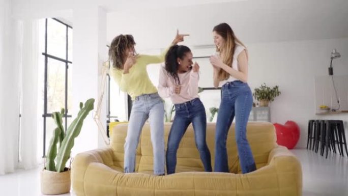 三个有趣的女性朋友站在室内的沙发上跳舞。多种族快乐的女人玩耍和庆祝快乐的女孩在家里一起享受快乐。