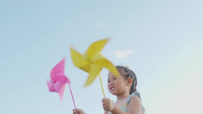 亚洲儿童女孩玩风车，快乐小女孩微笑在麦田里拿着小风轮或风车玩具在公园花园的夏日