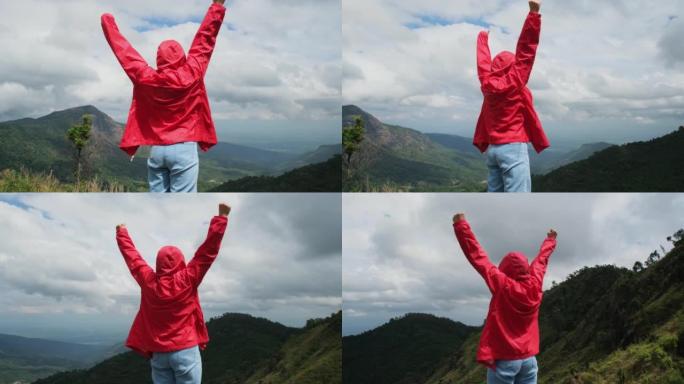 穿着红色雨衣的年轻亚洲徒步旅行者站在山顶，双臂高举，欣赏美景。旅程和成功概念