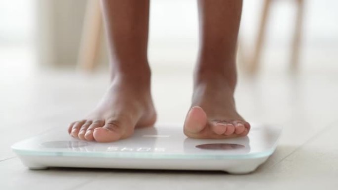 黑人女孩站在秤上测量体重的腿。非裔美国女性赤脚在家用体重秤。节食、控制和测量