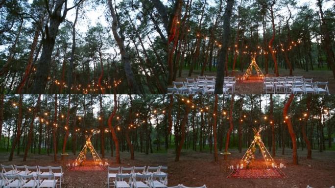 晚上松林户外婚礼场地波西米亚提皮拱门装饰上的节日串灯照明。夏季乡村婚礼上，复古串灯灯泡花环闪耀在椅子