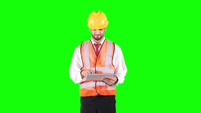 男性承包商戴着头盔并在绿色屏幕背景下站在工作室中使用数字平板电脑