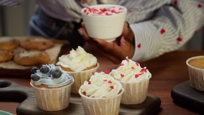 非裔美国厨师用浆果和五彩纸屑装饰纸杯蛋糕特写。女人糖果制造商展示烹饪松饼，上面放着奶油，美味的甜点。