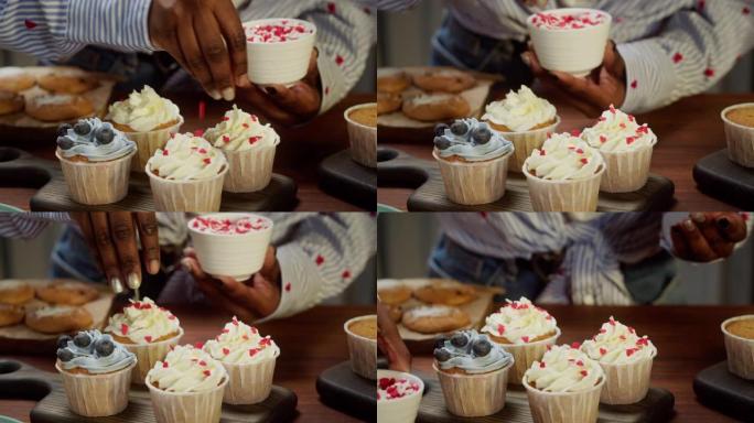 非裔美国厨师用浆果和五彩纸屑装饰纸杯蛋糕特写。女人糖果制造商展示烹饪松饼，上面放着奶油，美味的甜点。