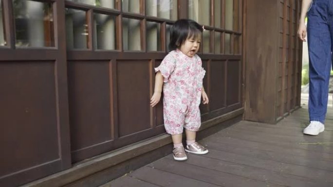 穿着和服的全长无辜的蹒跚学步的女孩正在移动，站在她的日本木屋旁，同时与走在附近的母亲玩耍