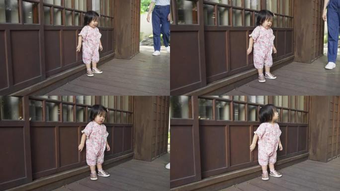 穿着和服的全长无辜的蹒跚学步的女孩正在移动，站在她的日本木屋旁，同时与走在附近的母亲玩耍