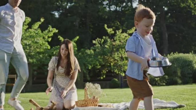 家庭野餐坐在院子里绿色的草地上，和一只小狗玩耍，快乐的笑儿子手里拿着碗